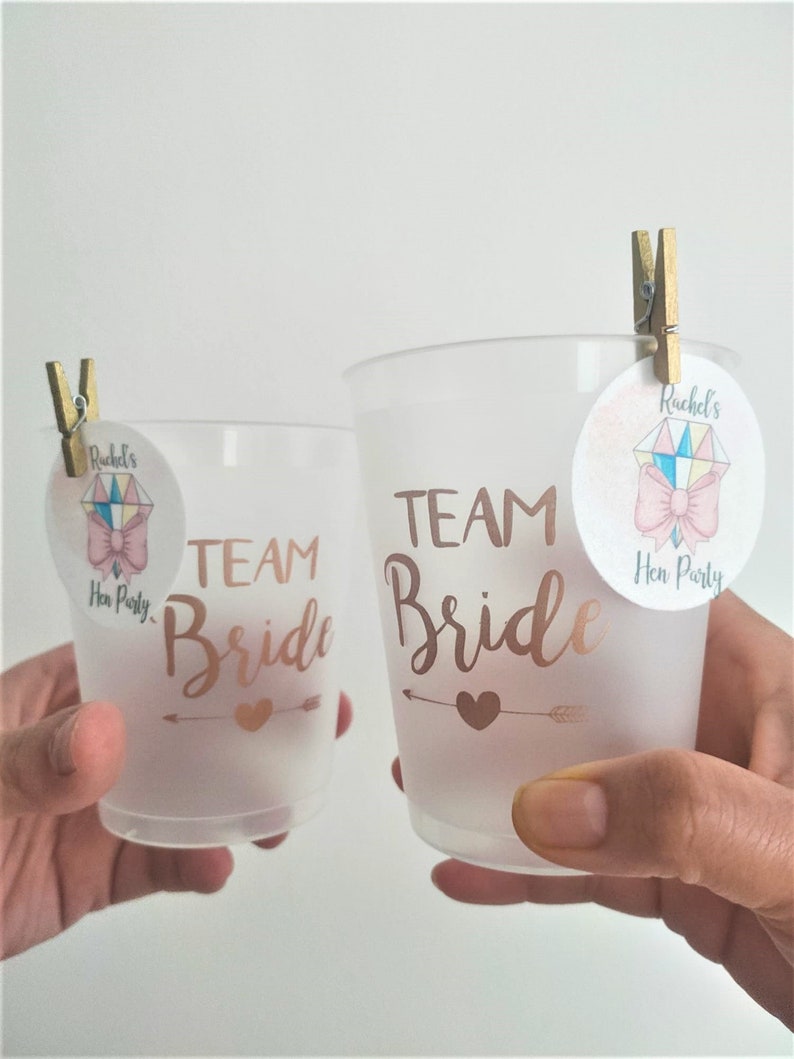 May include: Two frosted plastic cups with the text "Team Bride" in gold lettering. Each cup has a small pink and white sticker with a bow and the text "Rachel's Hen Party".