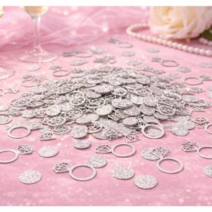 Peut inclure: Confettis argentés scintillants en forme de bagues de fiançailles et de cercles, dispersés sur une surface rose. Des verres à champagne et un collier de perles sont visibles en arrière-plan. Idéal pour les mariages et les célébrations.