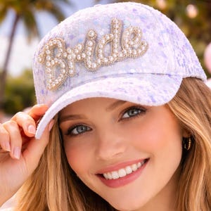 Puede incluir: Gorra de béisbol blanca con la palabra "Bride" escrita con adornos de perlas. La gorra tiene un patrón de lentejuelas de color púrpura claro. La imagen muestra a una persona que lleva la gorra, sonriendo, con un fondo tropical.