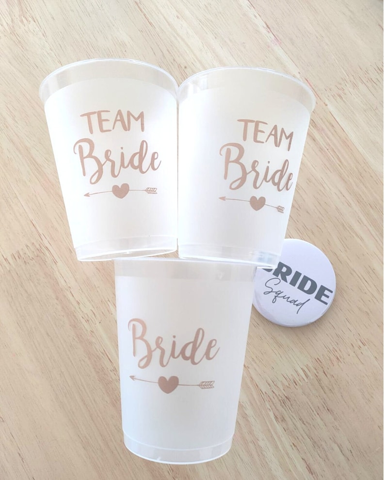 May include: Three white plastic cups with the text "Team Bride" in gold lettering and a gold arrow and heart design. A white button with the text "Bride Squad" in black lettering.