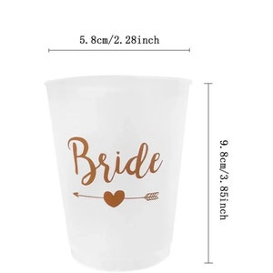 May include: White plastic cup with the word "Bride" in gold lettering and a gold arrow with a heart. The cup is 5.8 cm wide and 9.8 cm tall.