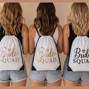 Peut inclure: Trois sacs à dos blancs avec des cordons noirs et les mots "Bride Squad" en or rose, or et noir. Chaque sac a un diamant au-dessus du texte. Les femmes portent des shorts en jean et des débardeurs noirs.