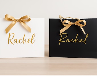 May include: Two gift bags, one white and one black, are displayed. Each bag has a gold ribbon bow at the top and the name "Rachel" in gold script. The black bag has rope handles.