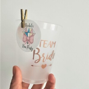 May include: A clear plastic cup with the text "Team Bride" in gold lettering and a small gold heart with an arrow pointing to the right. A small round sticker with the text "Rachel's Hen Party" and a pink bow is attached to the cup with a wooden peg.