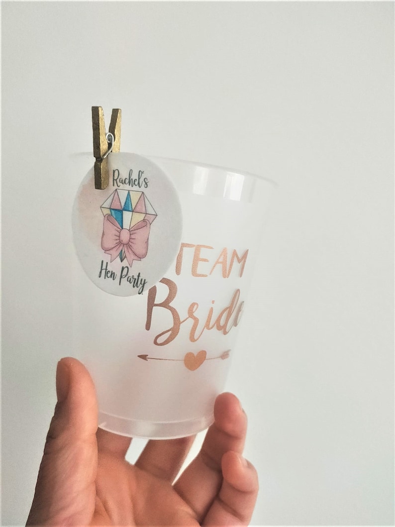 May include: A clear plastic cup with the text "Team Bride" in gold lettering and a small gold heart with an arrow pointing to the right. A small round sticker with the text "Rachel's Hen Party" and a pink bow is attached to the cup with a wooden peg.
