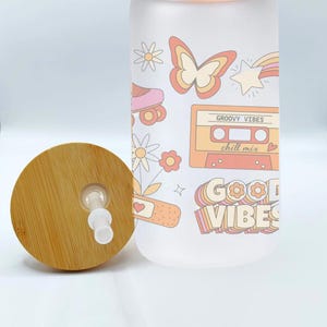 May include: A frosted glass water bottle with a bamboo lid and straw. The bottle has a retro design with a butterfly, roller skates, a cassette tape, and the text "Groovy Vibes chill mix" and "Good Vibes".