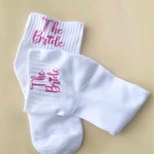 Peut inclure: Une paire de chaussettes blanches à la cheville avec l'inscription "The Bride" en lettres roses sur chaque chaussette. Les chaussettes ont un symbole de cœur rose entre les mots "The" et "Bride".