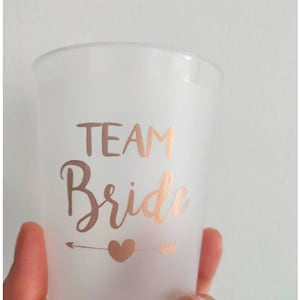 May include: A frosted plastic cup with a rose gold arrow and heart design. The cup says "Team Bride" in rose gold lettering.