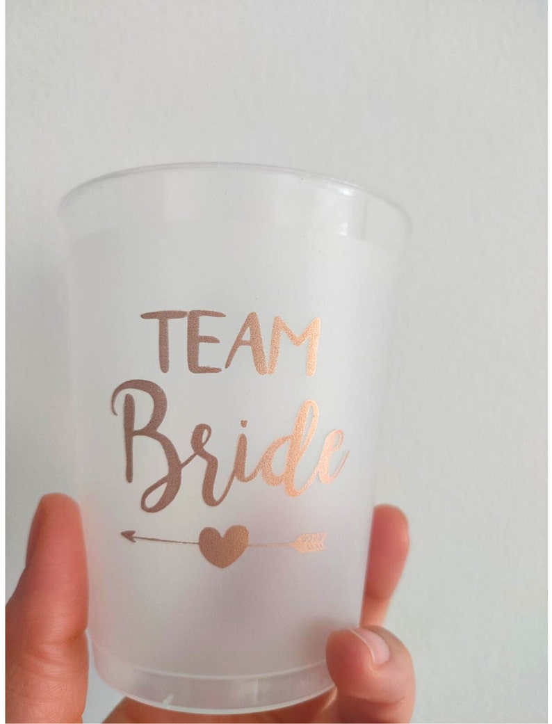 May include: A frosted plastic cup with a rose gold arrow and heart design. The cup says "Team Bride" in rose gold lettering.