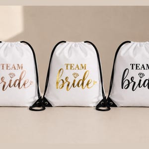 Peut inclure: Trois sacs blancs à cordon de serrage avec des cordons noirs. Chaque sac porte l'inscription "TEAM bride" en lettres métalliques or rose, or et noir. Un diamant est placé entre les mots.