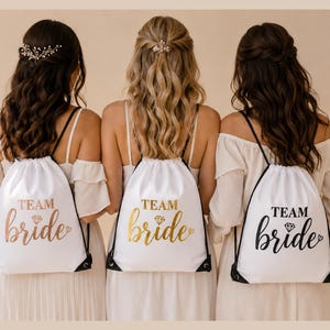 May include: Three women wearing white dresses and carrying white drawstring backpacks. The backpacks have the words "TEAM bride" printed on them in gold, black, and rose gold lettering. The women are standing in front of a neutral background.