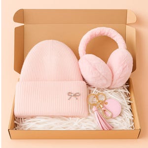 May include: A pink winter gift set in a cardboard box. The set includes a knit beanie with a bow accent, fluffy earmuffs, and a keychain with a tassel. The items are arranged on shredded paper.