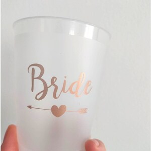May include: A frosted plastic cup with a rose gold arrow and heart design and the word "Bride" in rose gold lettering.