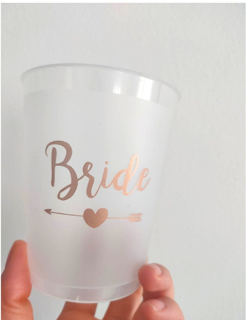 May include: A frosted plastic cup with a rose gold arrow and heart design and the word "Bride" in rose gold lettering.