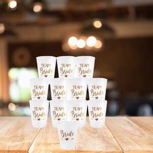 May include: A stack of white plastic cups with gold lettering that says "Team Bride". There is one cup at the bottom that says "Bride".
