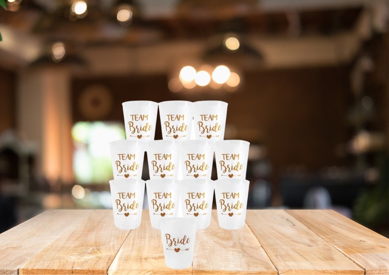 May include: A stack of white plastic cups with gold lettering that says "Team Bride". There is one cup at the bottom that says "Bride".