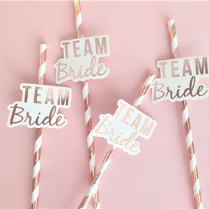 May include: Four pink and white striped paper straws with white paper flags that say "Team Bride" in rose gold foil lettering.