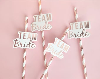 May include: Four pink and white striped paper straws with white paper flags that say "Team Bride" in rose gold foil lettering.