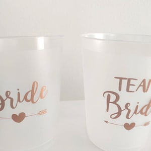 May include: Two white plastic cups with rose gold lettering. One cup says "Bride" with an arrow and heart. The other cup says "Team Bride" with an arrow and heart.
