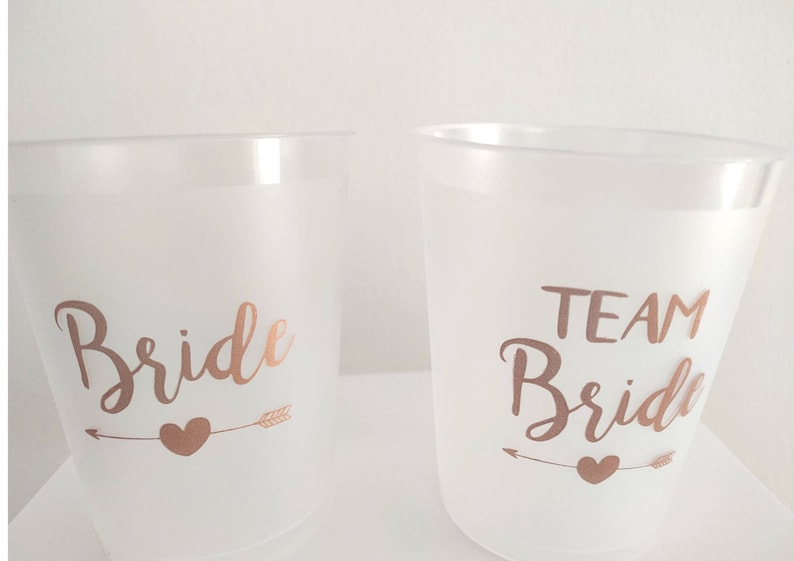 May include: Two white plastic cups with rose gold lettering. One cup says "Bride" with an arrow and heart. The other cup says "Team Bride" with an arrow and heart.