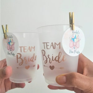 May include: Two clear plastic cups with the text "Team Bride" in gold lettering. Each cup has a small pink and gold circle sticker attached with a wooden peg. The stickers have a pink bow and the text "Rachel's Hen Party".