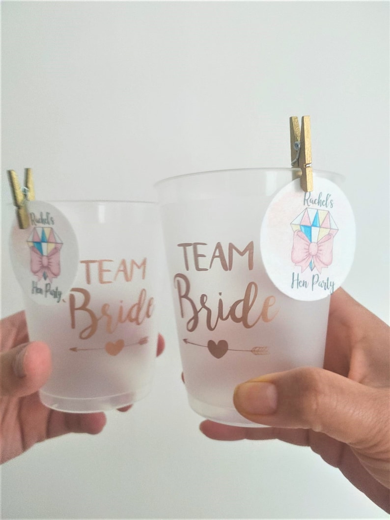 May include: Two clear plastic cups with the text "Team Bride" in gold lettering. Each cup has a small pink and gold circle sticker attached with a wooden peg. The stickers have a pink bow and the text "Rachel's Hen Party".