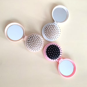 May include: Three compact, foldable pocket mirrors with a brush on one side and a mirror on the other. The mirrors are pink and white with black bristles.
