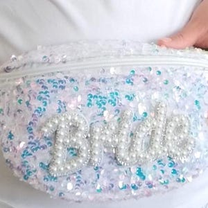 May include: A white sequined fanny pack with the word "Bride" spelled out in pearl beads. The bag has a zipper closure and a gold-colored clasp. The sequins are iridescent, reflecting shades of blue, pink, and white.