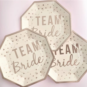 May include: Three white paper plates with a rose gold design. The plates have a scalloped edge and feature the text "Team Bride" with hearts and rings.