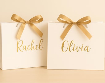 May include: Two white gift bags with gold bows and handles. The bags have gold lettering with the names "Rachel" and "Olivia". The bags are on a light beige surface.