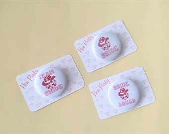 May include: Three white round buttons with pink text and a pink disco ball with a pink cowboy hat on top. The buttons say "Team Bride", "Bride" and "Bride Squad". The buttons are on white cards with a pink diamond pattern.