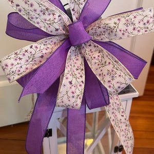 May include: A purple and white floral ribbon bow tied on a white lantern. The bow is made of two different ribbons, one with a white background and a purple floral pattern, and the other a solid purple ribbon.