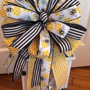 May include: A decorative bow featuring a bee theme. The bow has layers of ribbon with a honeycomb pattern, black and white stripes, and bee illustrations. The bow is yellow, white, and black.