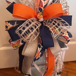 May include: A decorative bow featuring orange, navy, and beige ribbons. The bow has a layered design with burlap netting and ribbons printed with fall leaves. The bow is likely for seasonal decor, such as a lantern or wreath.