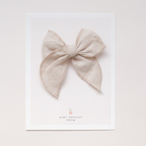 May include: A beige fabric bow with a stitched edge. The bow is displayed on a white card with the text "Mini Apricot Company".