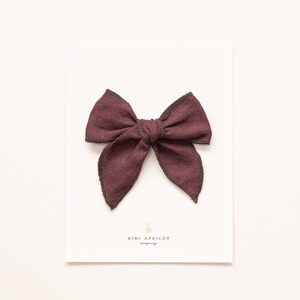 May include: A burgundy fabric bow with a brown stitched edge. The bow is on a white card with the text "MINI APRICOT" printed on it.