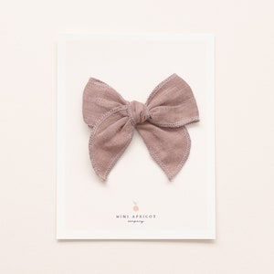 May include: A dusty rose coloured fabric bow with a simple knot in the centre. The bow is on a white card with the text "mini apricot company" printed on it.