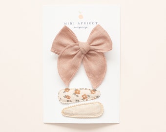 May include: A set of three hair clips featuring a blush pink bow, a floral print clip, and a textured clip. The clips are displayed on a white card with the text "MINI APRICOT company".