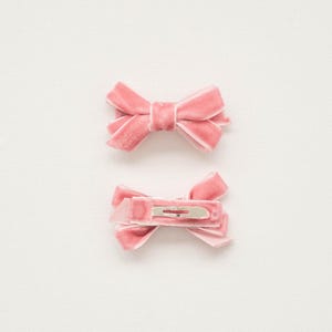 May include: Two pink velvet hair bows. One bow is displayed from the front, and the other shows the back with a silver metal clip. The bows are a soft, muted pink color and are set against a white background.