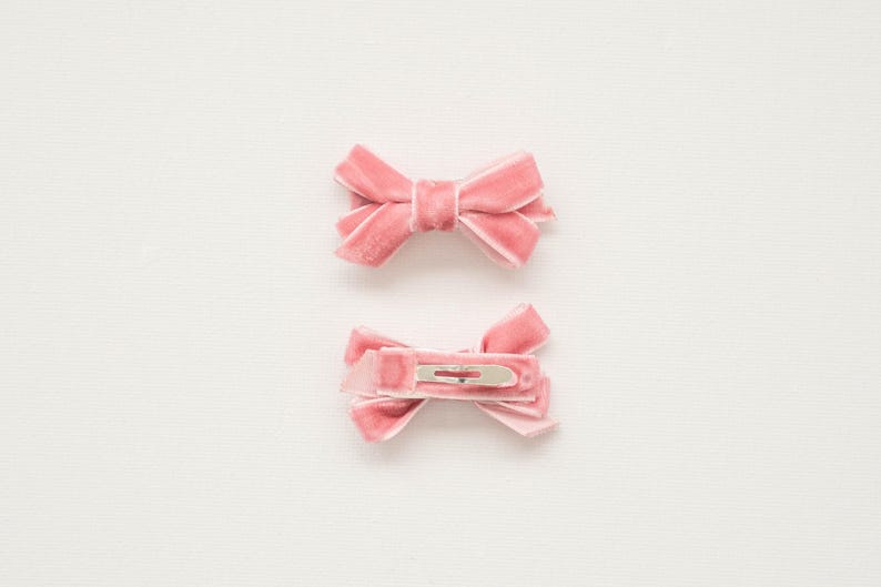 May include: Two pink velvet hair bows. One bow is displayed from the front, and the other shows the back with a silver metal clip. The bows are a soft, muted pink color and are set against a white background.