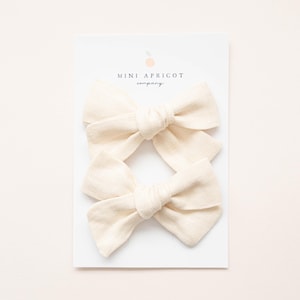 Puede incluir: Dos lazos para el pelo de tela color crema sobre una tarjeta blanca. Los lazos están atados en el centro y tienen un diseño simple y clásico. La tarjeta tiene el texto "MINI APRICOT company" en una fuente delicada. El fondo es de un color suave y neutro.
