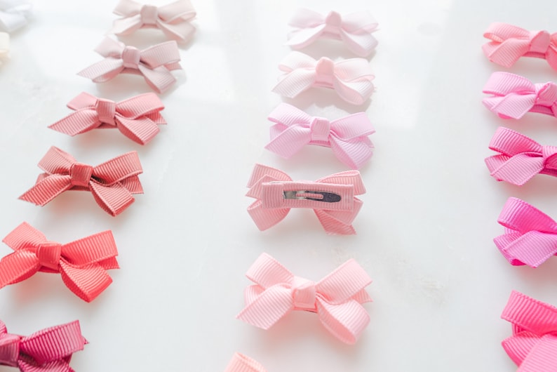 May include: Rows of small, pink hair bows in various shades, from pale blush to vibrant fuchsia. Each bow is made of ribbed ribbon and attached to a metal clip, perfect for accessorizing hairstyles.