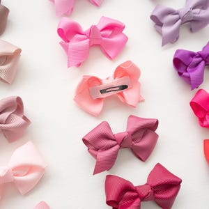 May include: An assortment of colorful hair bows in shades of pink, purple, and brown. The bows are made of a ribbed fabric and are arranged on a white surface. Some bows have metal clips on the back.