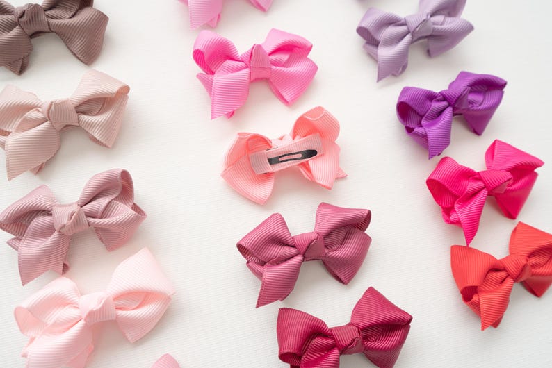 May include: An assortment of colorful hair bows in shades of pink, purple, and brown. The bows are made of a ribbed fabric and are arranged on a white surface. Some bows have metal clips on the back.