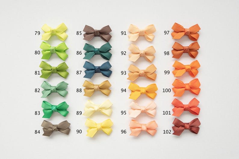 May include: A collection of small, colorful bows in various shades, including yellow, green, blue, and orange. Each bow is labeled with a number from 79 to 102. These bows are likely used for crafting or hair accessories.