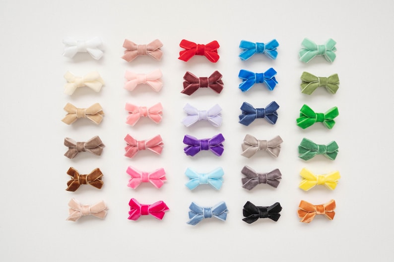 May include: A collection of small, velvet bow hair accessories in various colors, including white, pink, red, blue, green, purple, and brown. The bows are arranged on a white surface.