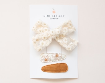 May include: A set of three hair clips featuring a white floral bow and two smaller clips with a floral pattern and a solid brown fabric. The clips are displayed on a white card with the text "MINI APRICOT company".