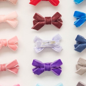 May include: An assortment of velvet hair bows in various colors, including pink, red, blue, purple, and gray. Each bow is neatly arranged and features a classic bow shape with a metal clip on the back. These accessories are suitable for all ages.