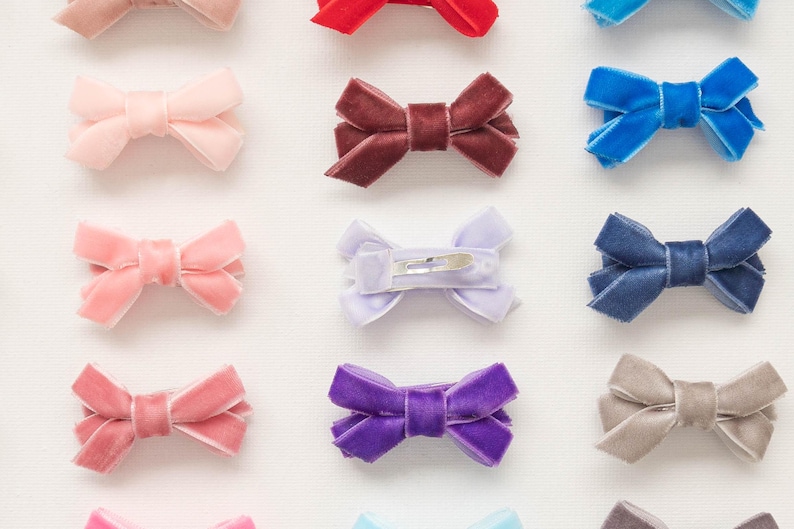 May include: An assortment of velvet hair bows in various colors, including pink, red, blue, purple, and gray. Each bow is neatly arranged and features a classic bow shape with a metal clip on the back. These accessories are suitable for all ages.