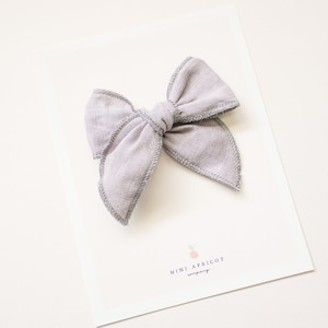 May include: A light gray fabric bow with a gray stitched edge. The bow is on a white card with the text "Mini Apricot Company" printed on it.